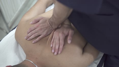 A-chiropractor-cracks-the-upper-middle-back-portion-of-a-young-adult-caucasian-male-patient-as-he-lies-topless-face-down-on-a-massage-table-in-a-physiotherapy-clinic,-closeup-top-down-view