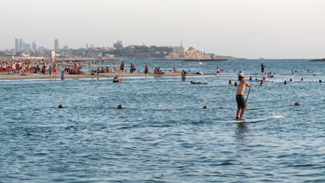 Hombre-Haciendo-Paddle-Surf-En-La-Playa-De-Tel-Aviv-Con-Gente-Nadando-En-El-Fondo-En-Israel