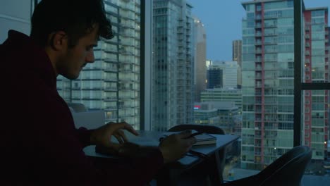 Man-writing-a-letter-or-writing-in-his-journal-in-his-apartment,-with-the-city-view-in-the-background