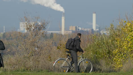 Junger-Mann-Steigt-Auf-Fahrrad-Mit-Industriedämpfen-Im-Hintergrund,-Nachhaltiger-Transport