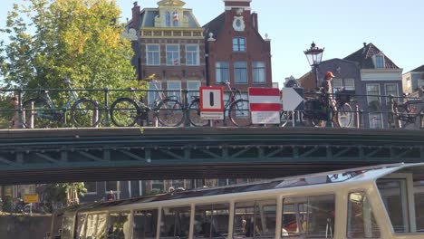Un-Barco-Turístico-Conduciendo-Lentamente-Bajo-El-Puente-Peatonal-Sobre-El-Canal-En-Amsterdam