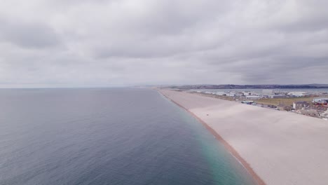 Drone-shot-flying-backwards-along-Chesil-beach-on-a-sunny-day,-Weymouth,-Dorset,-UK