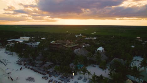 Aerial-Slow-Orbit-View-Of-Beach-Huts-And-Resorts-On-Akiin-Beach-In-Tulum-With-Orange-Sunset-Skies-In-Background