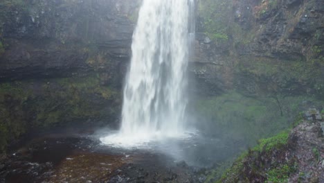 Aufsteigende-Luftdrohnenaufnahme-Eines-Dramatischen-Wasserfalls-Mit-Höhle-Dahinter-Und-Geschnittenen-Baumstämmen-4k