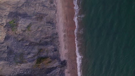 Top-down-drone-shot-moving-upwards-of-green-sea's-waves-and-cliffs-on-the-Jurassic-Coast,-Dorset,-UK