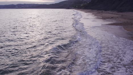 Slow-motion-drone-shot-moving-backwards-of-waves-crashing-on-the-beach,-Jurassic-Coast,-Dorset,-UK