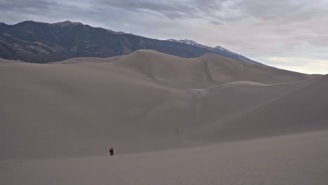 Female-hiker-walks-towards-tent-set-up-in-middle-of-dune-field-with-mountains