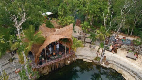 Aerial-view-of-a-tropical-wooden-building-next-to-a-cenote,-Tulum,-Mexico,-surrounded-by-palm-trees,-with-people-relaxing