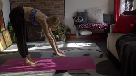 Beautiful-skinny-fit-long-blonde-haired-caucasian-white-girl-or-women-in-yoga-outfit-stretching-and-doing-upward-dog-pose-on-pink-yoga-mat-and-stretching-on-rug-in-front-of-apartment-fireplace