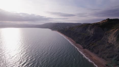 High-drone-shot-moving-backwards-along-the-coast-showing-the-cliffs-and-beach-on-the-Jurassic-Coast,-Dorset,-UK