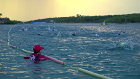 Slow-motion-wide-shot-of-a-lifeguard-cheering-up-the-competitors-of-a-triathlon-swimming-in-the-sea-at-dawn