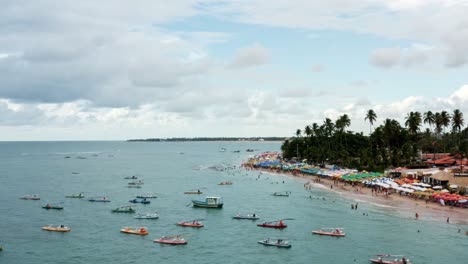 Left-trucking-aerial-drone-shot-of-the-Porto-de-Galinhas-or-Chicken-Port-beach-with-anchored-sailboats,-colorful-umbrellas,-and-tourists-swimming-in-the-crystal-clear-ocean-water-in-Pernambuco,-Brazil