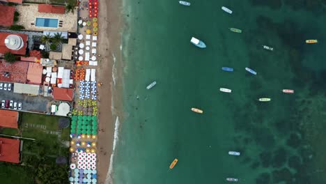 Bird's-eye-top-down-moving-aerial-shot-of-the-famous-Porto-de-Galinhas-or-Chicken-Port-beach-in-Pernambuco,-Brazil-with-colorful-umbrellas,-tourists-swimming-in-the-natural-pools,-and-sailboats