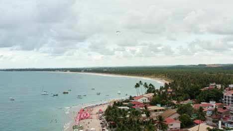 Descending-aerial-drone-wide-shot-of-the-Porto-de-Galinhas-or-Chicken-Port-beach-with-anchored-sailboats,-a-hang-glider,-and-tourists-swimming-in-the-crystal-clear-ocean-water-in-Pernambuco,-Brazil