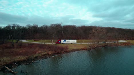 pull-away-aerial-of-a-red-semi-truck-and-white-trailer-parked-on-side-of-a-lake