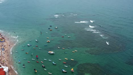 Descending-aerial-drone-wide-shot-of-the-Porto-de-Galinhas-or-Chicken-Port-beach-with-anchored-sailboats-and-tourists-swimming-in-the-crystal-clear-ocean-water-in-Pernambuco,-Brazil