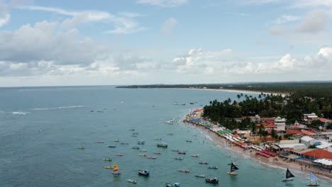 Descending-aerial-drone-wide-shot-of-the-Porto-de-Galinhas-or-Chicken-Port-beach-with-anchored-sailboats-and-tourists-swimming-in-the-crystal-clear-ocean-water-in-Pernambuco,-Brazil