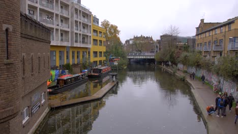 High-angle-shot-over-Regent's-canal-in-Camden-area-in-London,-UK-with-locals-passing-by-beside-the-canal-on-a-cloudy-day