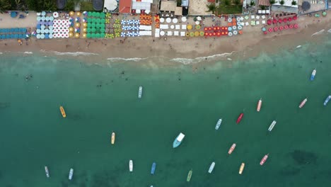 Bird's-eye-top-down-aerial-moving-shot-of-the-famous-Porto-de-Galinhas-or-Chicken-Port-beach-in-Pernambuco,-Brazil-with-hotels,-colorful-umbrellas,-tourists-swimming-in-the-natural-pools-and-sailboats