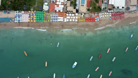 Bird's-eye-top-down-aerial-shot-of-the-famous-Porto-de-Galinhas-or-Chicken-Port-beach-in-Pernambuco,-Brazil-with-hotels,-colorful-umbrellas,-tourists-swimming-in-the-natural-pools,-and-sailboats