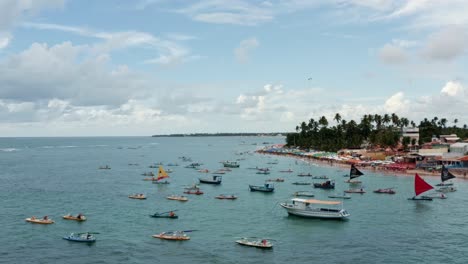 Dolly-in-aerial-drone-wide-shot-of-the-Porto-de-Galinhas-or-Chicken-Port-beach-with-anchored-sailboats,-a-hang-glider,-and-tourists-swimming-in-the-crystal-clear-ocean-water-in-Pernambuco,-Brazil