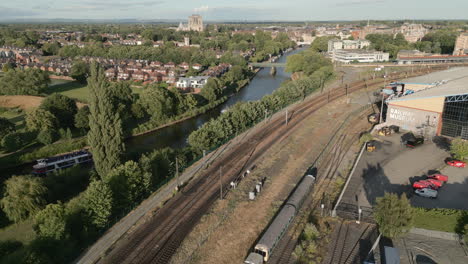 Aerial-Drone-Shot-Along-York-Railway-Line-and-River-Ouse-with-Tourist-Ferry-Passing-by-and-York-Minster-Cathedral-in-Background-on-Sunny-Evening-Sunset-North-Yorkshire-United-Kingdom