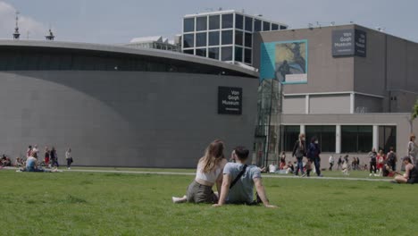 General-slow-motion-shot-of-a-couple-sitting-on-the-grass-looking-at-the-Van-Gogh-Museum,-located-in-Amsterdam,-The-Netherlands