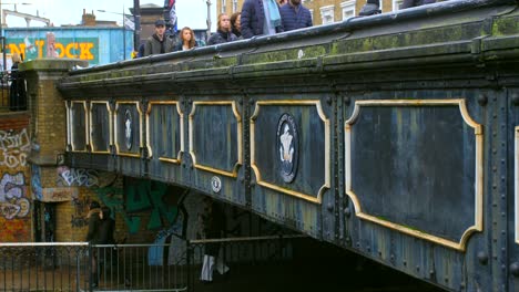 Gente-Cruzando-El-Puente-Histórico-Sobre-El-Canal-De-Los-Regentes-En-El-Mercado-De-Camden-Lock-En-Londres,-Reino-Unido