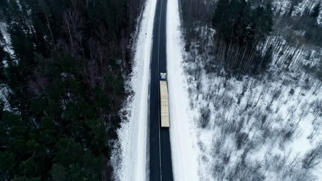 Aerial-View-Of-Loaded-Truck-Driving-On-The-Road-Through-Snowy-Pine-Forest-In-Winter