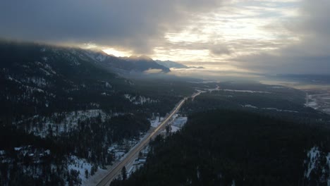 Splendid-Aerial-Approach-Shot-of-Cariboo-Highway-95-Running-into-the-Horizon-Surrounded-by-Grand-Mountains-Range-Covered-in-Clouds-During-Warm-Sunrise-Light:-Panoramic-View-of-Winter-Landscape