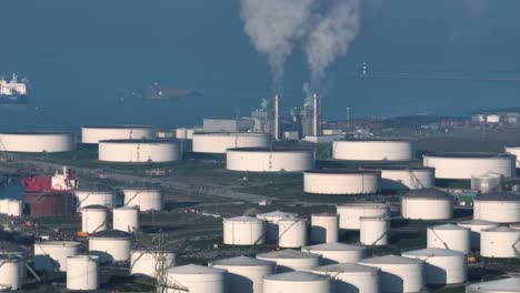 Wide-aerial-view-of-large-capacity-oil-storage-tanks-near-the-ocean-with-ships-in-the-background-and-industrial-facility-with-stacks-emitting-steam