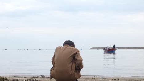 View-From-Behind-Young-Pakistani-Boy-Sat-In-Squat-Position-Looking-Out-To-Sea-From-Gwadar-Beach