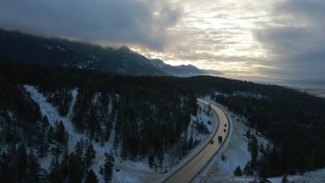 Early-Morning-Sunrise-Aerial-Reveal-of-Cars-Travelling-Along-Snow-Covered-Mountain-Cariboo-Highway-95-with-Reflections-on-Road-in-Mystical,-Cloudy-Scene-in-British-Columbia,-Canada