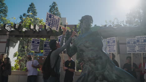 A-Young-Woman-Academic-Worker-puts-a-UAW-on-Strike-Sign-in-the-Hands-of-a-Statue-on-UCLA's-Campus
