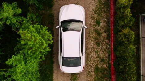 Medium-Wide-Top-Down-Aerial-Drone-Shot-of-White-Car-Driving-Down-Dirt-Driveway-into-a-Car-Garage-attached-to-a-Home