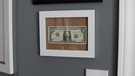 A-one-dollar-bill-hung-on-a-cork-board-with-a-glass-front-panel-and-white-wood-frame-showing-of-financial-accomplishment-in-somebody's-bedroom