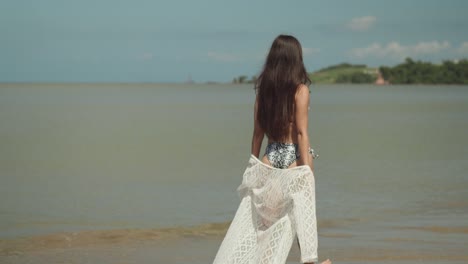 Mujer-Joven-Caminando-Por-La-Playa-En-Bikini-Y-Olas-Oceánicas-Y-Cielos-Azules-En-El-Fondo