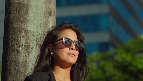 Erstaunliche-Gesichtsnahaufnahme-Einer-Latina-Frau-Mit-Sonnenbrille-An-Einem-Sonnigen-Tag-Mit-Hohen-Gebäuden-Im-Hintergrund