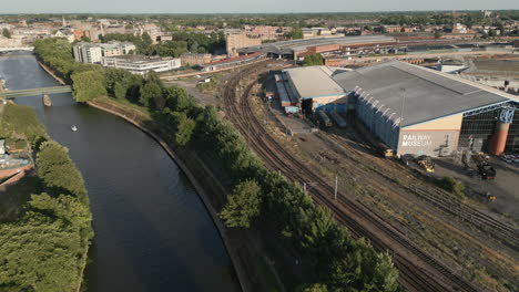 Aerial-Drone-Shot-of-National-Railway-Museum-and-River-Ouse-with-Boat-Travelling-down-River-with-York-Railway-Station-in-View-on-Sunny-Evening---York-City-North-Yorkshire-United-Kingdom
