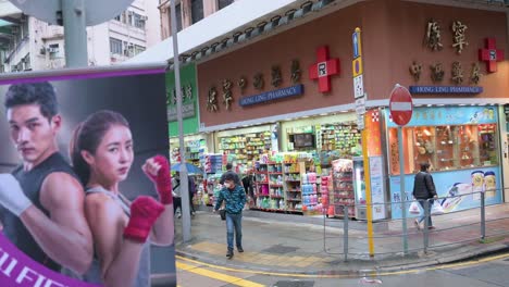 Chinese-pedestrians-walk-through-a-zebra-crossing-in-front-of-a-pharmacy-offering-and-selling-personal-care,-hygiene-products,-and-drugs-during-flu-season-in-Hong-Kong