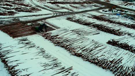 Aerial-reveal-shot-of-snow-covered-log-piles-and-heavy-equipment-at-sawmill-in-winter