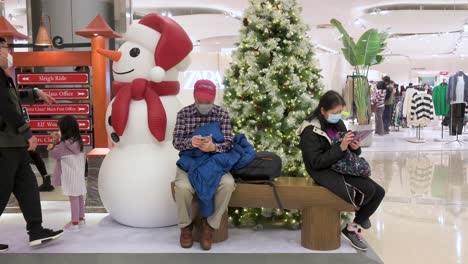 Chinesische-Käufer-Sitzen-Auf-Einer-Bank-Neben-Einem-Weihnachtsbaum-Und-Einer-Schneemanninstallation-In-Einem-Einkaufszentrum,-Während-Andere-Kunden-In-Hongkong-An-Ihnen-Vorbeigehen