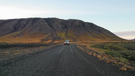 Aerial-Shot-Of-White-Land-Rover-Suv-Driving-On-Black-Icelandic-Road-With-Stunning-Black-And-Yellow-Mountain-Background-During-Adventure-Travel-Road-Trip