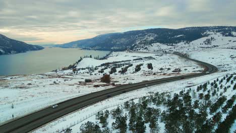 Winterwunderland-Im-Zentrum-Von-Okanagan:-Eine-Luftperspektive-Von-Autos,-Die-Auf-Dem-Highway-97-Inmitten-Einer-Verschneiten-Landschaft-Und-Eines-Waldsees-Fahren