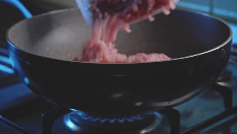 Side-view-close-up-of-raw-ground-or-minced-meat-getting-added-into-a-deep-nonstick-frying-skillet-pan-heating-up-on-a-clean-blue-flame-from-a-builtin-hob-cooktop-gas-stove