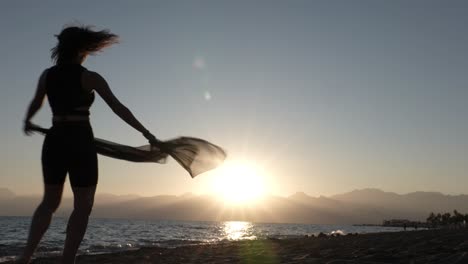 Silhouette-of-Young-Woman-in-Bikini-With-Shawl-Dancing-and-Spinning-on-Beach-With-Sunset-Above-Sea