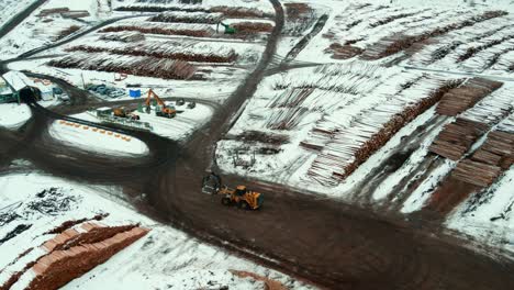 Aerial-view-of-wheel-loader-with-grapple-bucket-driving-through-large-piles-of-logs-covered-in-snow-at-Canadian-sawmill