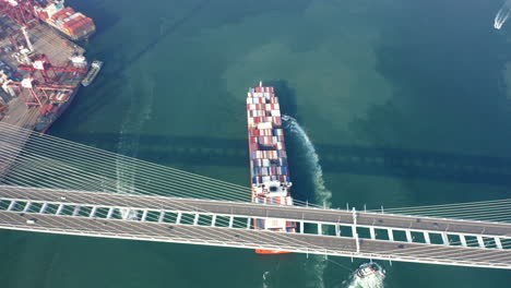 Rotes-Containerschiff,-Das-Von-Einem-Schlepp--Und-Schubboot-Unterstützt-Wird,-Das-Zum-Liegeplatz-An-Einem-Containerterminal-Navigiert,-Während-Es-Die-Steinmetzbrücke-Passiert