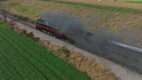 Drone-Parallel-Then-Behind-View-of-a-Steam-Passenger-Train-Blowing-Smoke-and-Steam-on-a-Sunny-Fall-Day