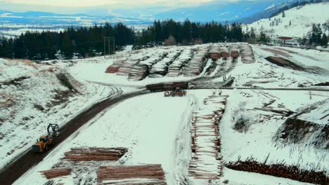Aerial-follow-shot-of-wheel-loader-with-grapple-bucket-maneuvering-towards-log-truck-to-unload-logs-at-Canadian-sawmill-in-winter